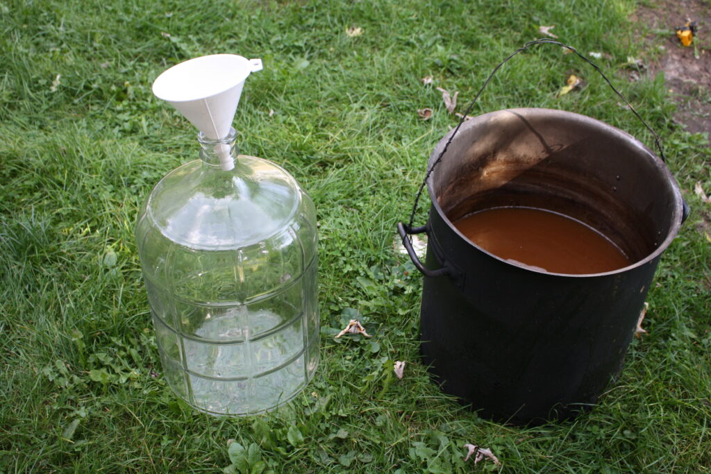 a glass carboy with a funnel next to a 10 gallon metal pot with beer wort.