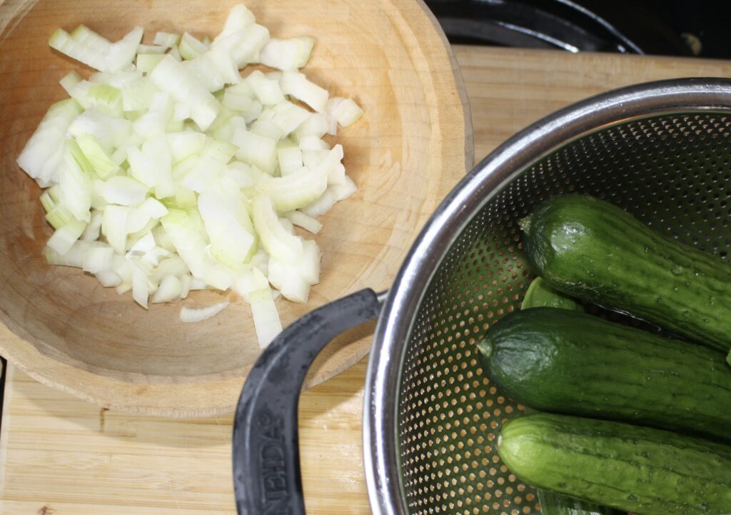 Cucumbers in a colander and minced onions in a wooden bowl on a cutting board