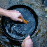 a cast-iron skillet being cleaned in a running stream with a twig scrubber