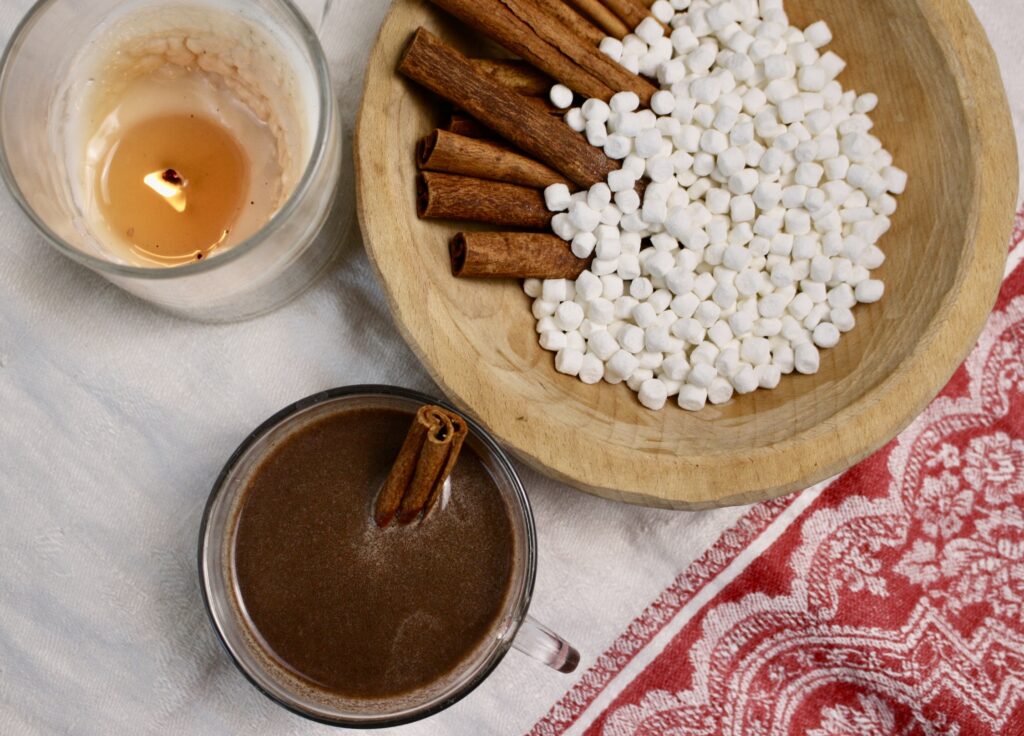 cup of hot cocoa sitting on Swedish cloth next to a candle and bowl of marshmallows and cinnamon sticks