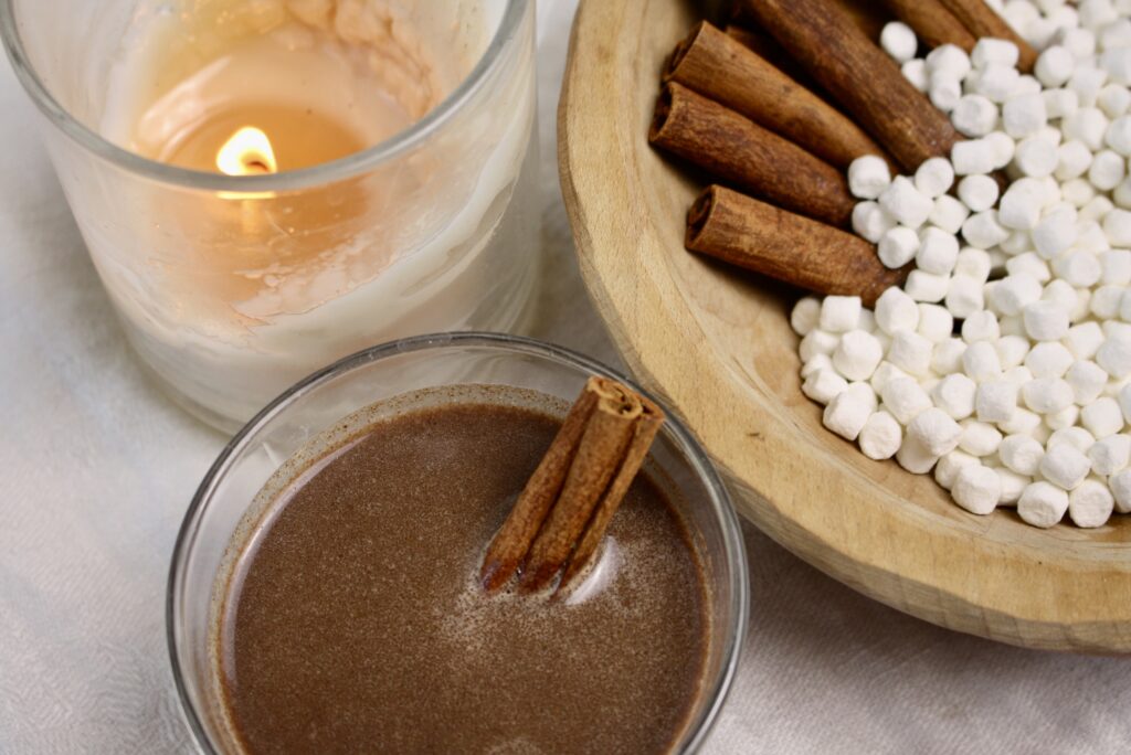 cup of hot cocoa next to a candle and bowl of cinnamon sticks and marshmallows