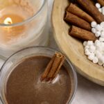 cup of hot cocoa next to a candle and bowl of cinnamon sticks and marshmallows