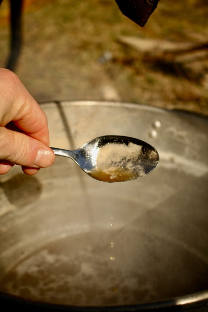 hand holding a metal spoon with maple syrup over a pot