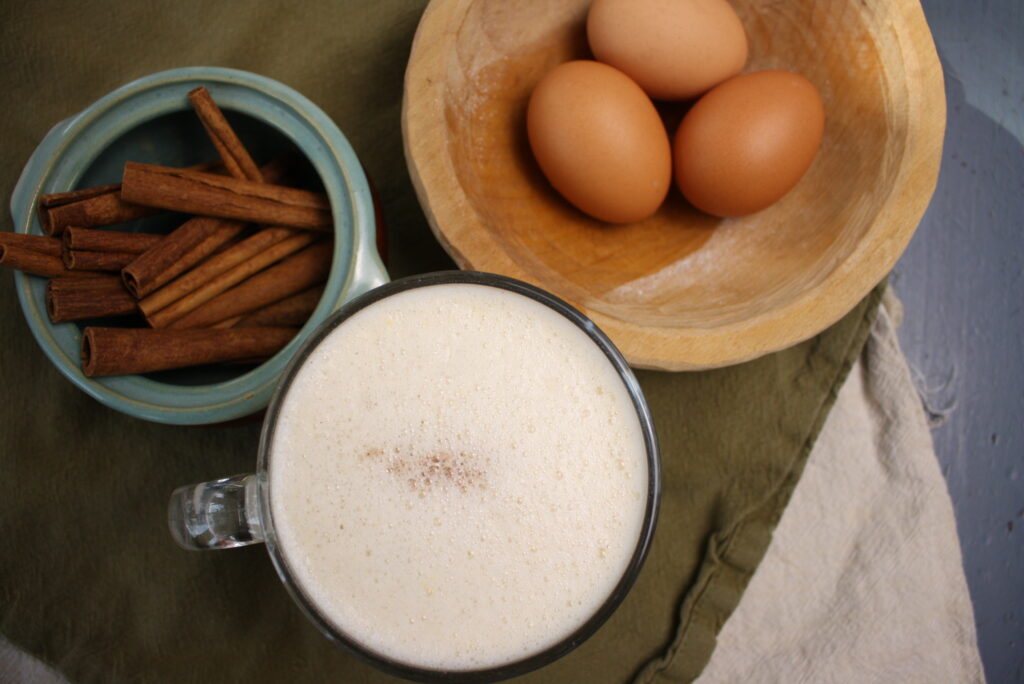 Caudle in a mug next to a bowl of cinnamon sticks and a bowl of eggs