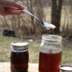 hand holding a metal spoon pouring maple syrup into a jar, next another jar on a wooden platter