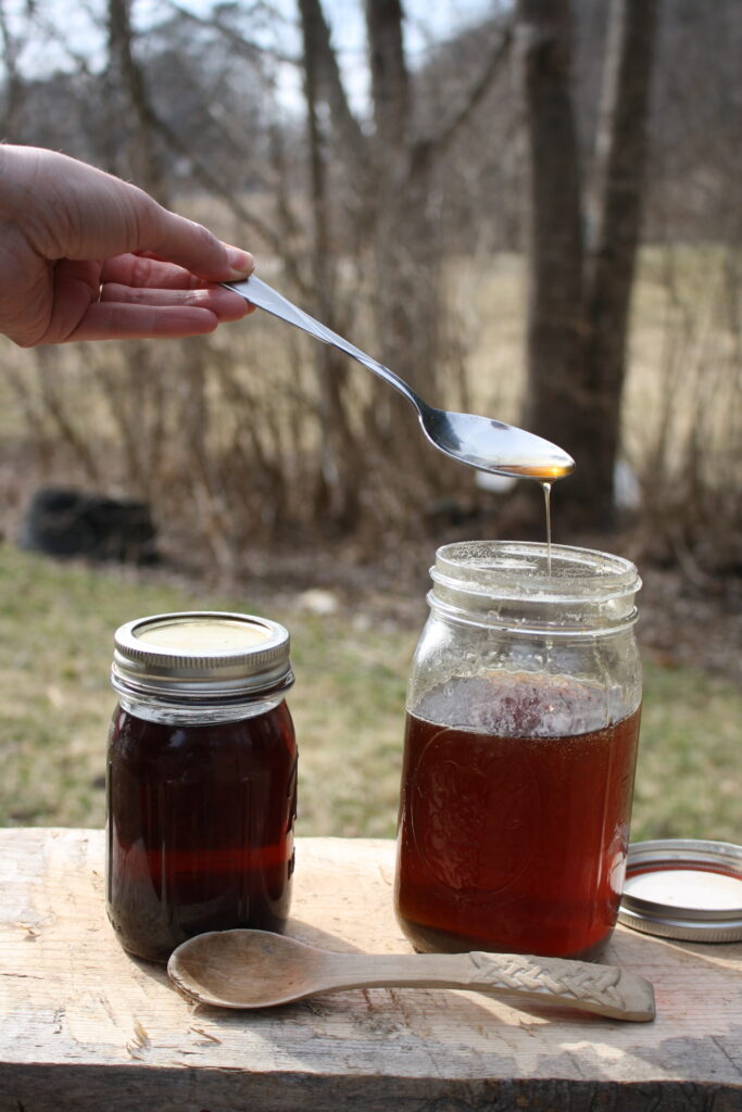 hand holding a metal spoon pouring maple syrup into a jar, next another jar on a wooden platter