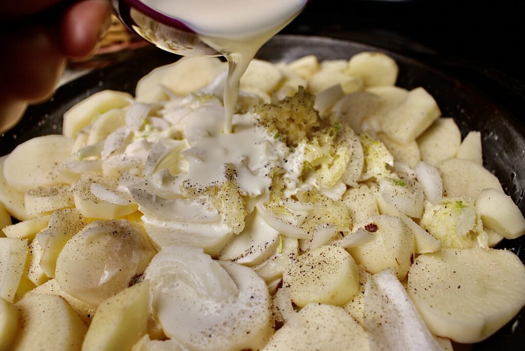 cream being poured over sliced potatoes with onions and garlic