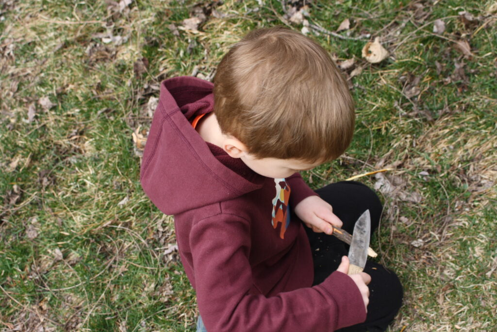 boy in a red sweater whittling with a stick with a knife