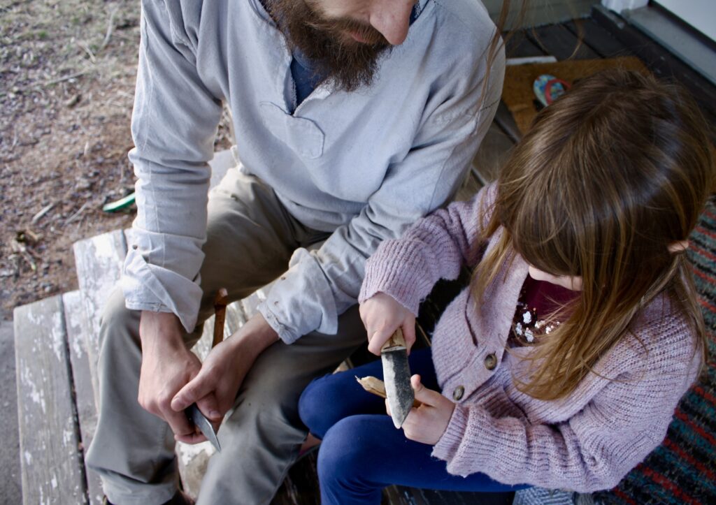 man sitting next to a girl whittling with a knife