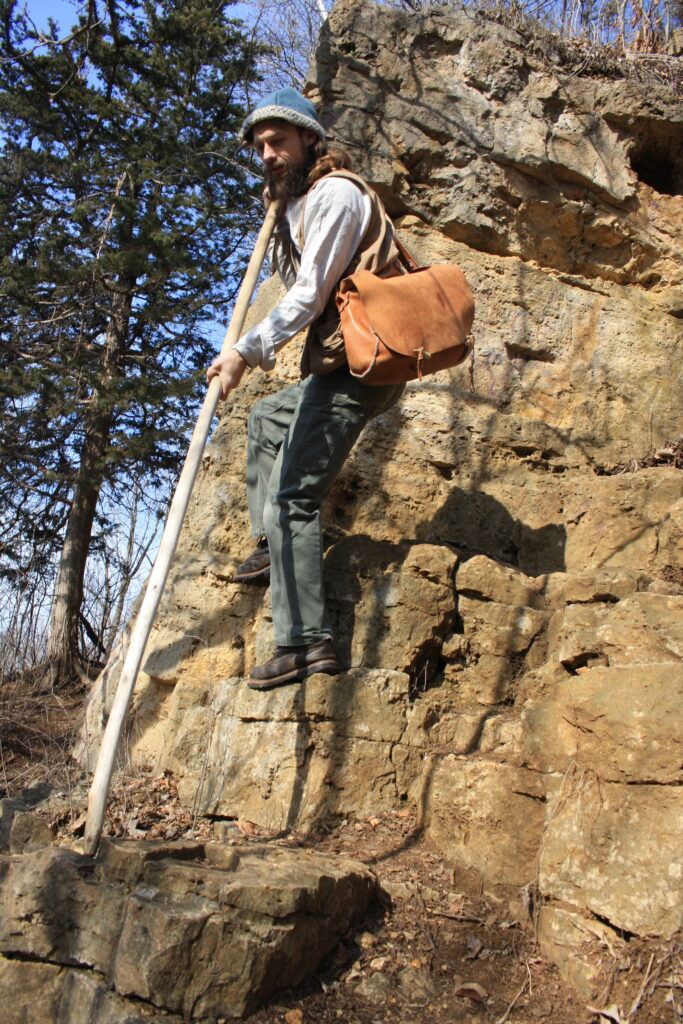 man on a boulder wearing a leather satchel and holding a long hiking stick