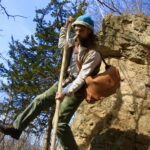 Man wearing a leather satchel leaping from a rock face with a long hiking stick