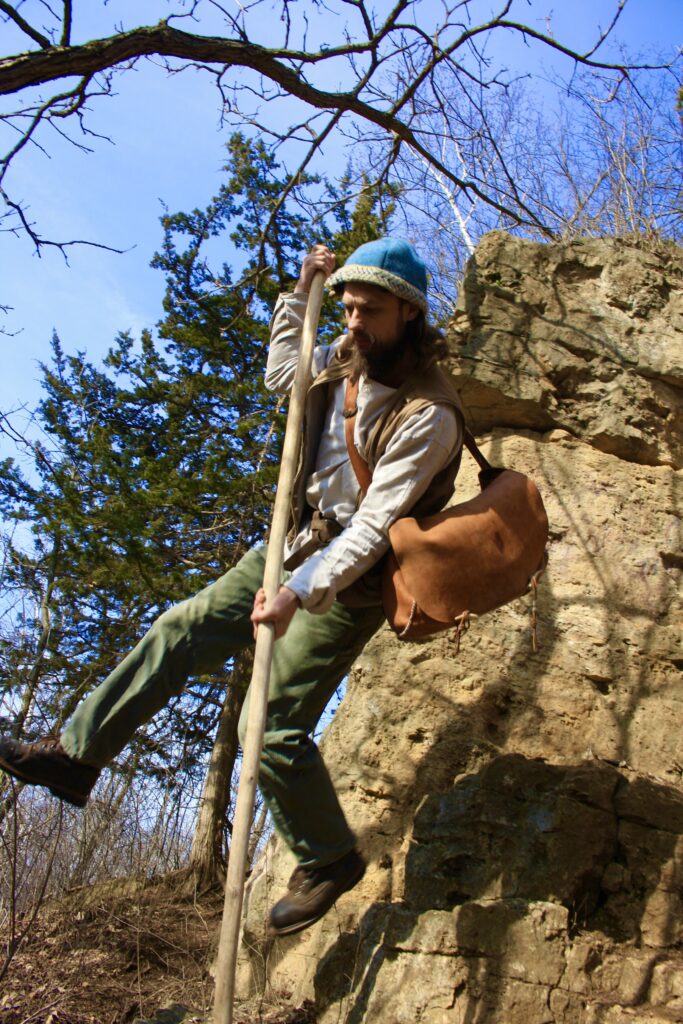 Man wearing a leather satchel leaping from a rock face with a long hiking stick