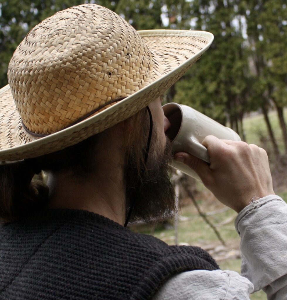 man wearing a straw hat in front of cedar trees drinking from a stoneware beer mug