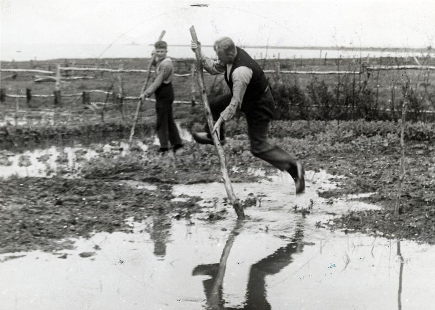 two dutch farmers in a black and white photo jumping over water with long poles