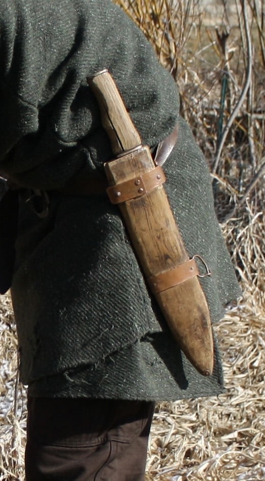 a machete hanging on a belt of a man wearing a long green coat