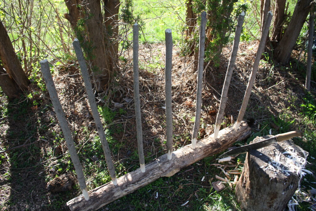 seven evenly-spaced posts stuck vertically into a log to make a wattle hurdle with a stump and a machete in the fore-ground