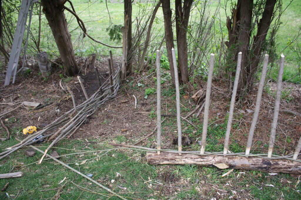 upright posts in a form with willow withes being woven into them to make a hurdle next to a small fence made by children