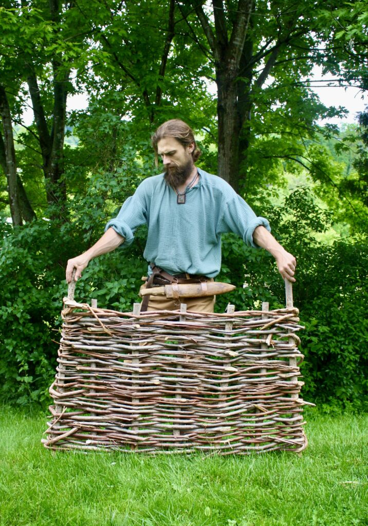 bearded man wearing a bush-knife, holding up a wattle hurdle