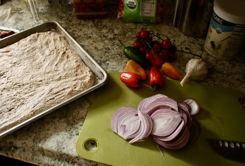 plain sourdough focaccia on a sheet pan and vegetables waiting to be chopped on a cutting board