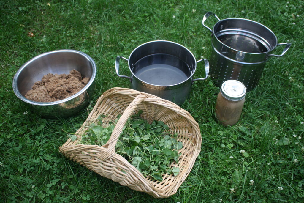 Supplies for making gruit--A bowl of spent grains, a basket of ale hoof, a pot of water, a metal strainer and a stare of ale-yeast sitting on the ground