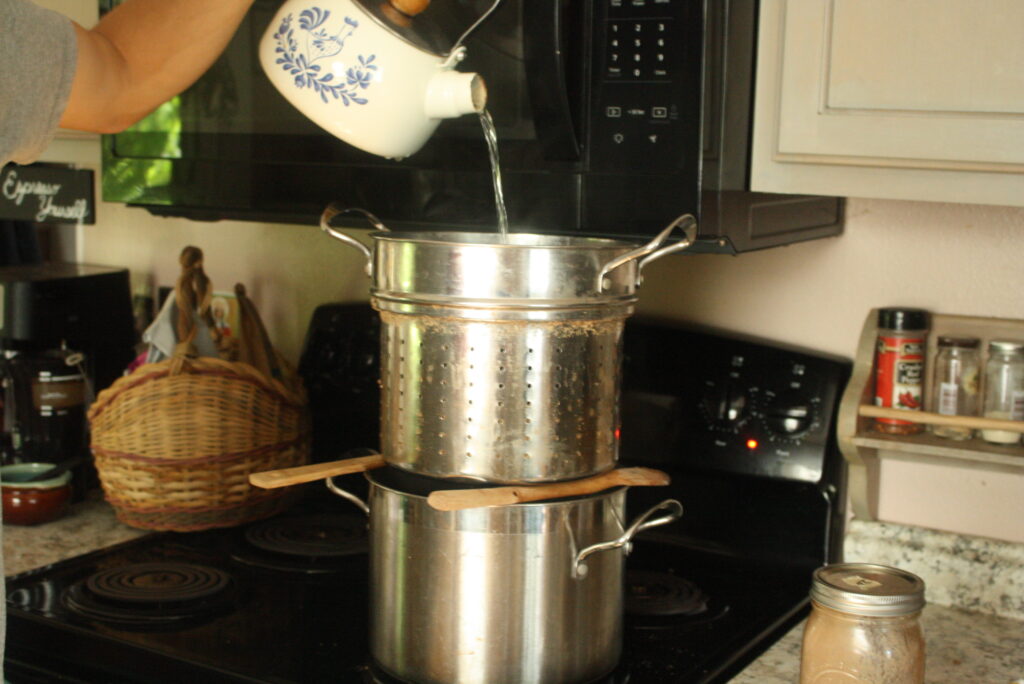 hot water from a tea-pot being poured over grains in a strainer over a metal pot 