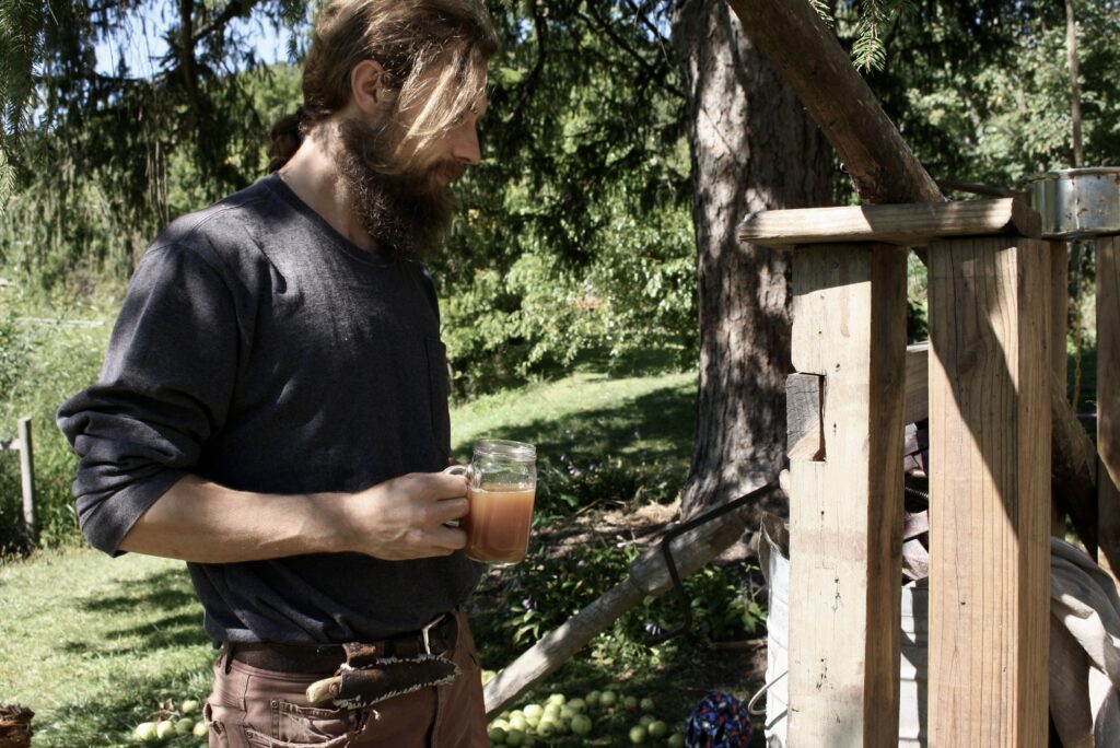 man holding a glass of apple cider next to a cider press under a spruce tree