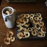 a tray of dry apple slices next to a ceramic jar filled with apple slices sitting on a wooden table