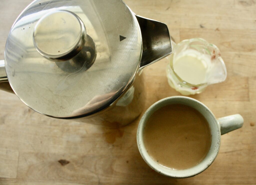 French press sitting on table by cup of coffee and cream