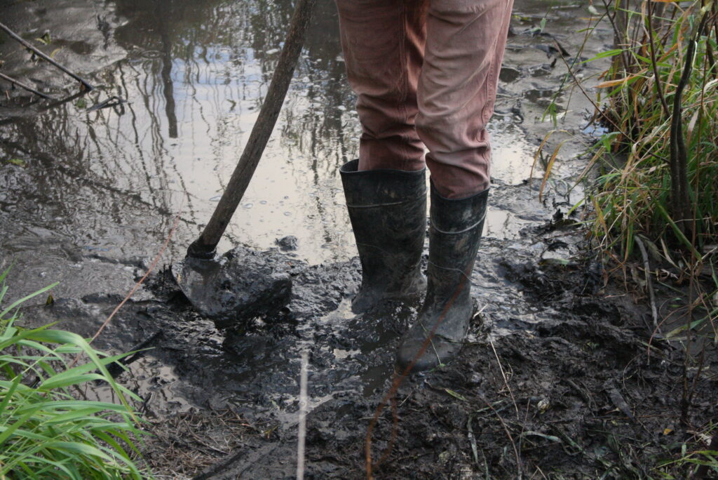 photo of persons feet in rubber boots standing in mud with a broad-hoe