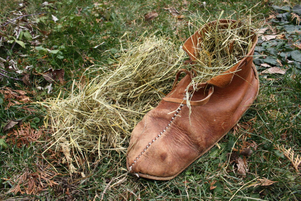 a leather shoe sitting on grass, stuffed with hay next to a small pile of hay