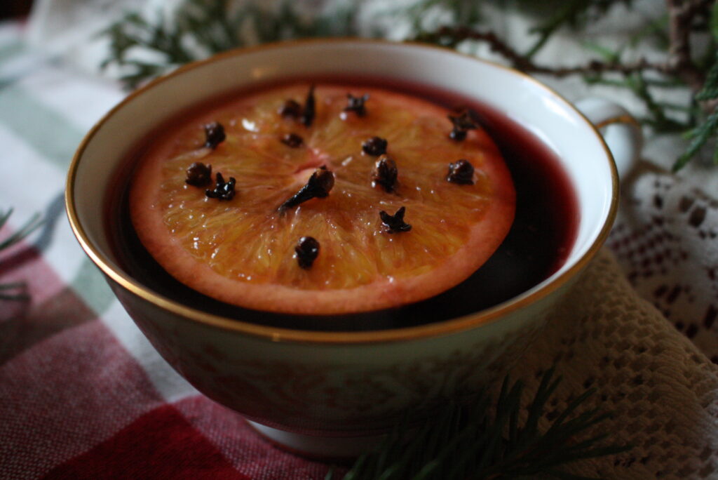 a mug of mulled wine on a plaid cloth next to evergreen boughs with an orange slice and cloves floating in it.