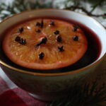 a mug of mulled wine on a plaid cloth next to evergreen boughs with an orange slice and cloves floating in it.