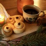 photo of traditional St Lucy's Feast food and drink. a candle, St Lucy buns, a mug of mulled wine, ginger bread and almonds on a wooden serving board