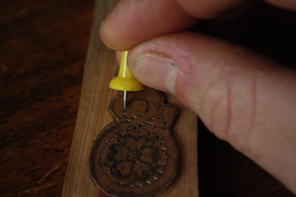 a hand holding a thumb tack, pushing holes through a cardboard template for an earring into the wood beneath