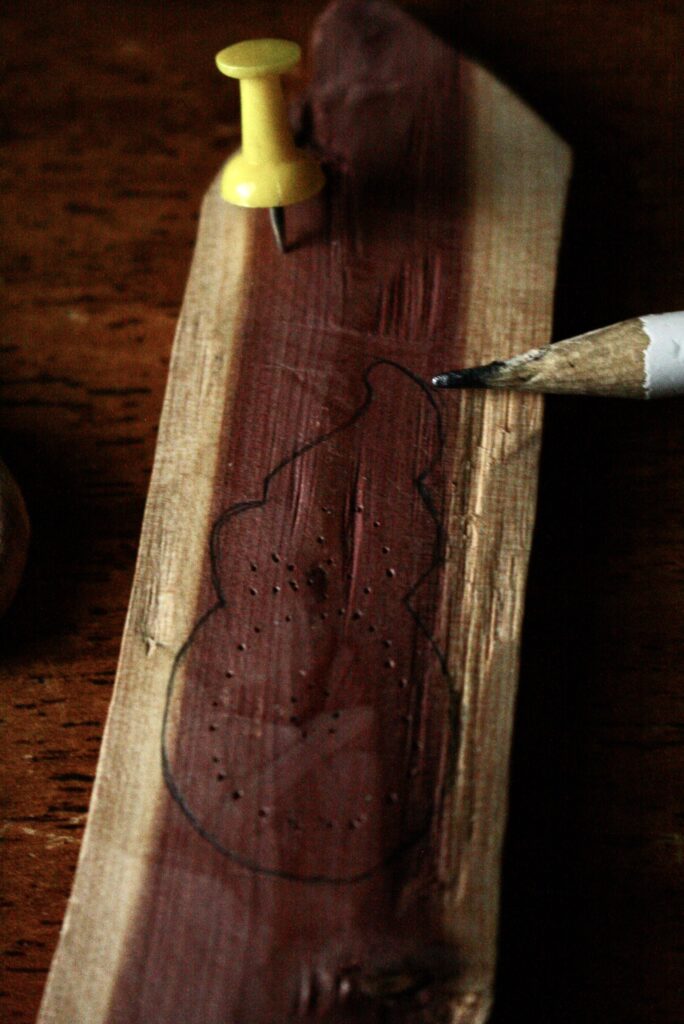 piece of wood with a thumbtack and an pencil, showing the traced shape for and pattern of holes to be carved out