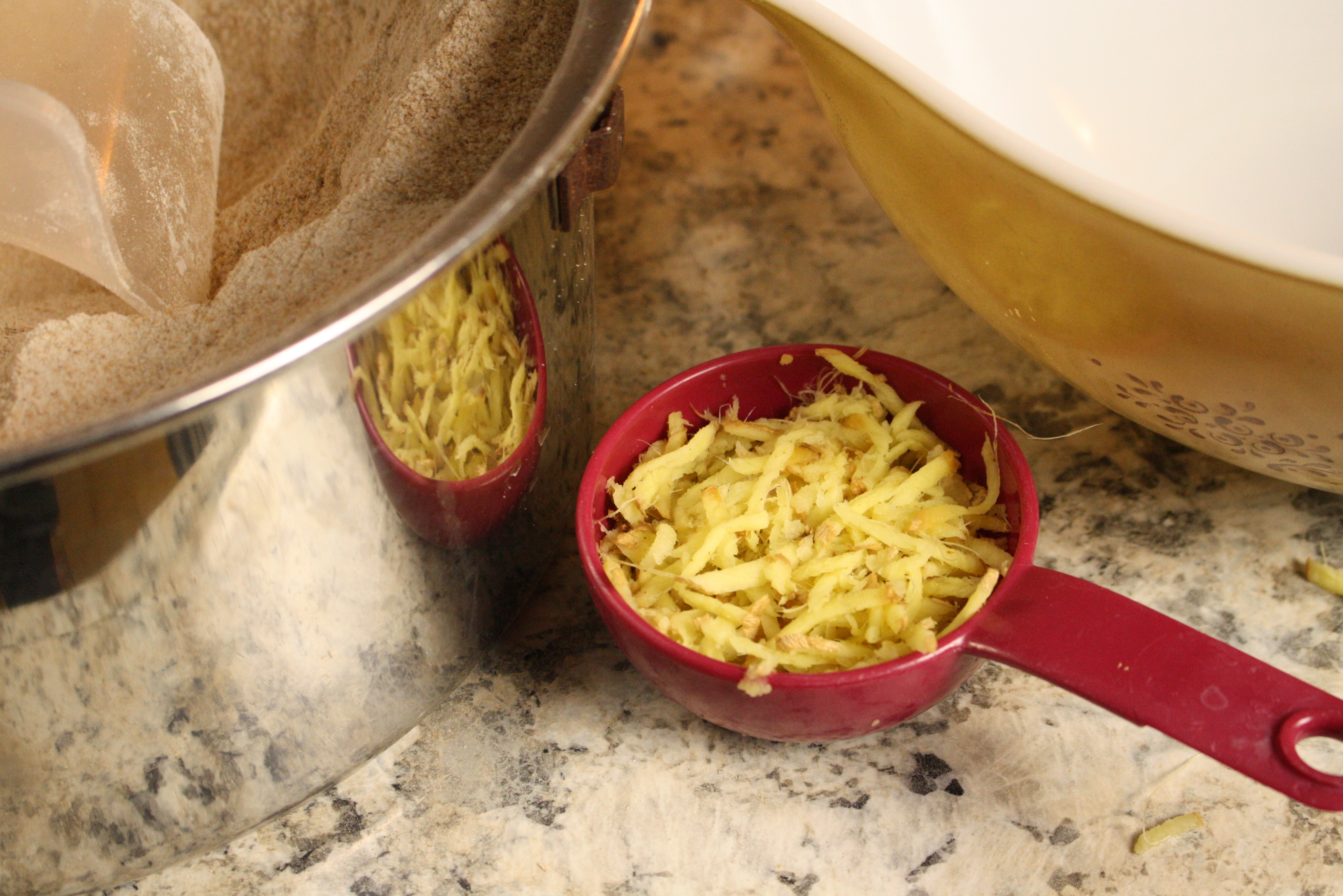 grated ginger in a red measuring cup next to a bowl and a stainless steel bowl with freshly milled flour