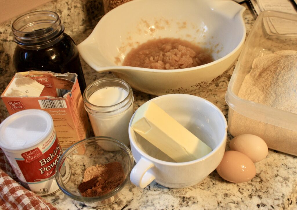 maple syrup, baking powder, baking soda, spices, milk, butter, applesauce, eggs, and milled flour on a countertop