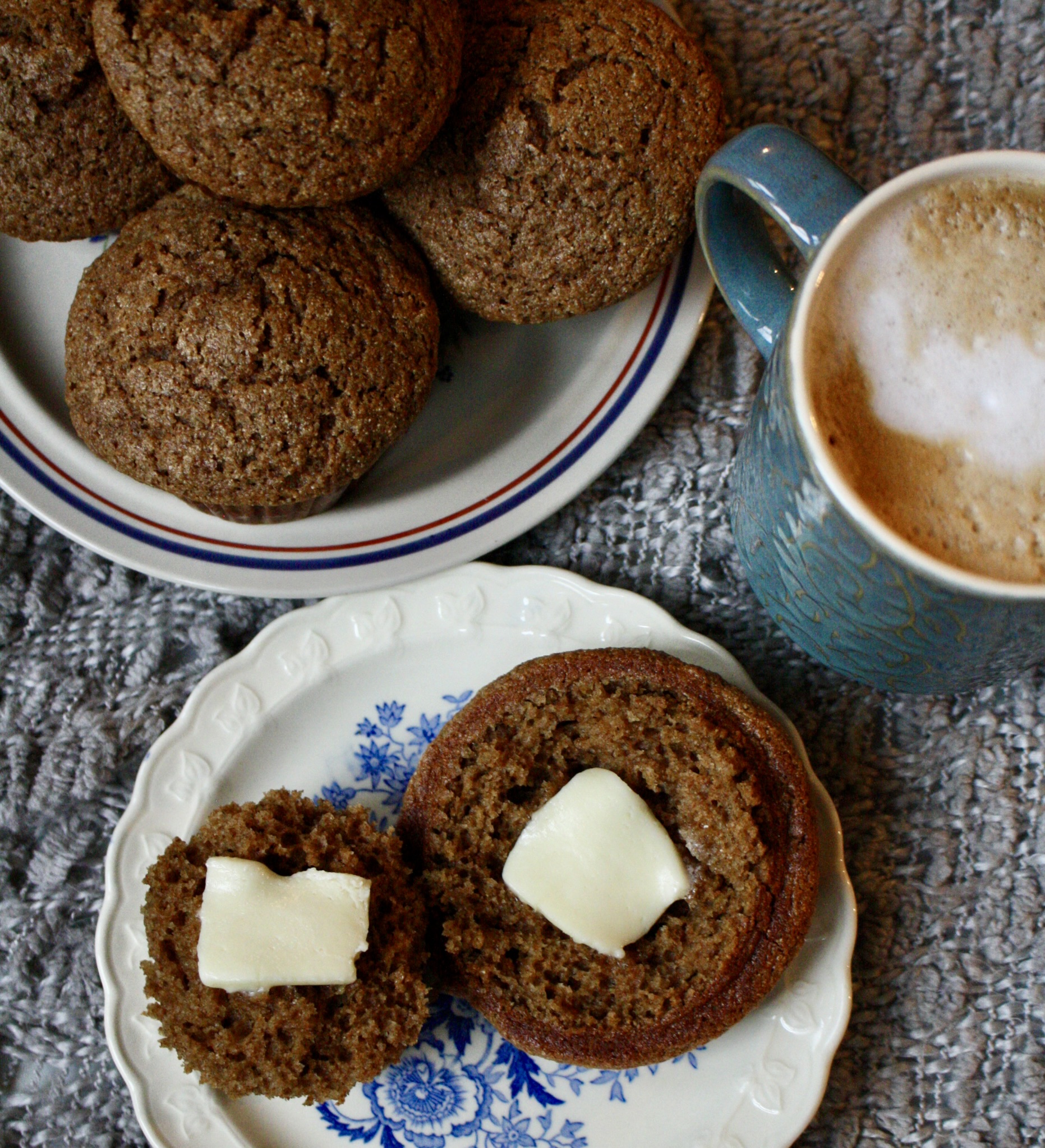 a spiced applesauce muffin cut in half with butter on each side sitting on a plate next to a cup of coffee and a plate full of muffins.