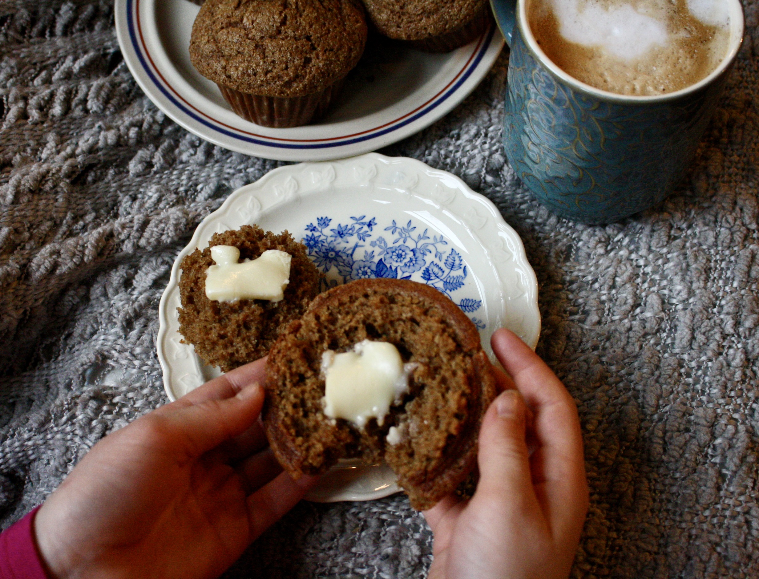 hands holding a muffin top with butter melting on it next to a latte and a plate of muffins