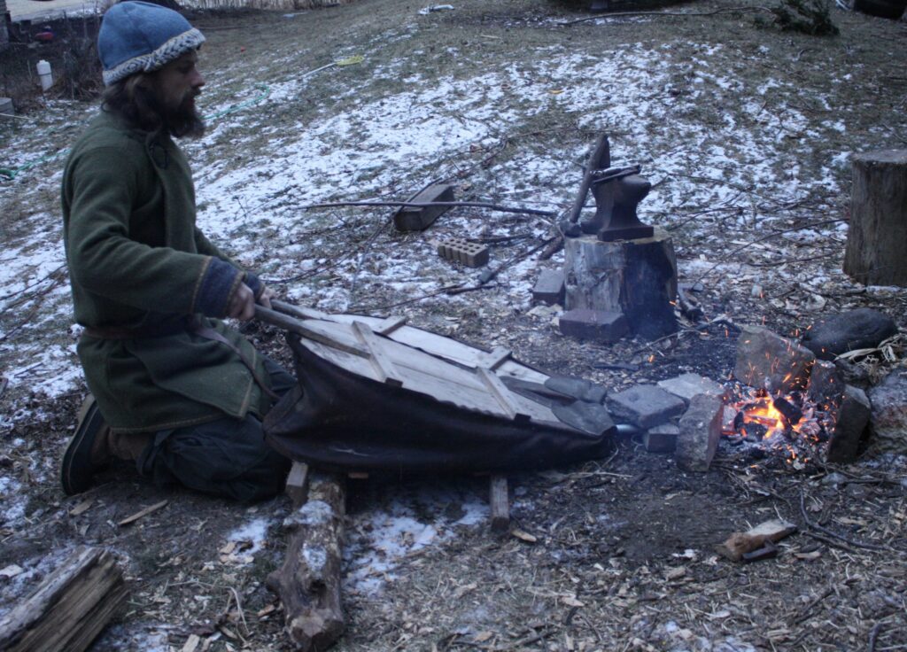 a man kneeling on the ground pumping a bellows for a forge fire set up on the ground