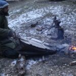 a man kneeling on the ground pumping a bellows for a forge fire set up on the ground