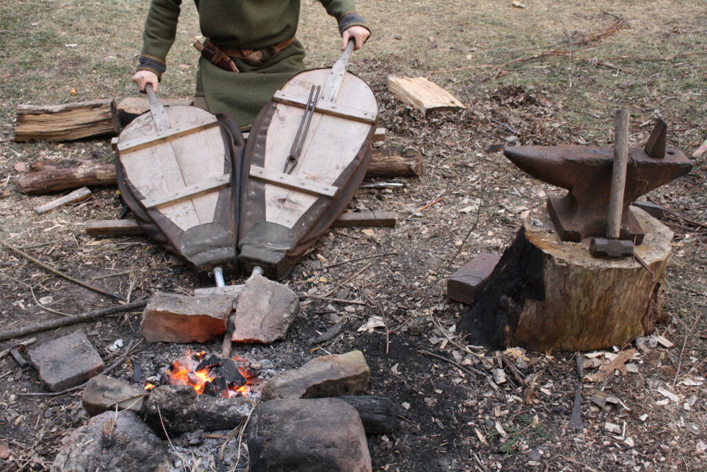 a man pumping two bellows on a backyard forge fire next to a stump with an anvil, hammer and tongs