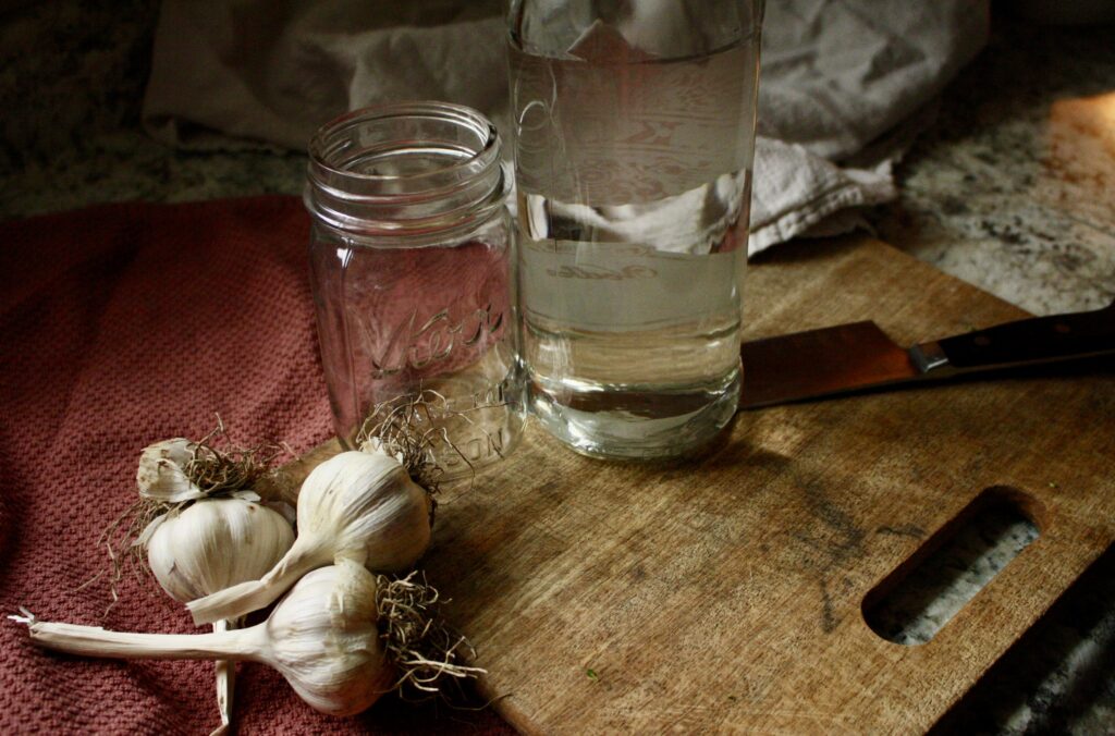 garlic bulbs, empty jar, and bottle of vodka sitting next to a cutting board