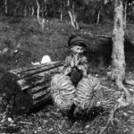 a black and white photo of a Sami boy holding two rolls of "shoe hay" standing next to an overturned wooden sledge.