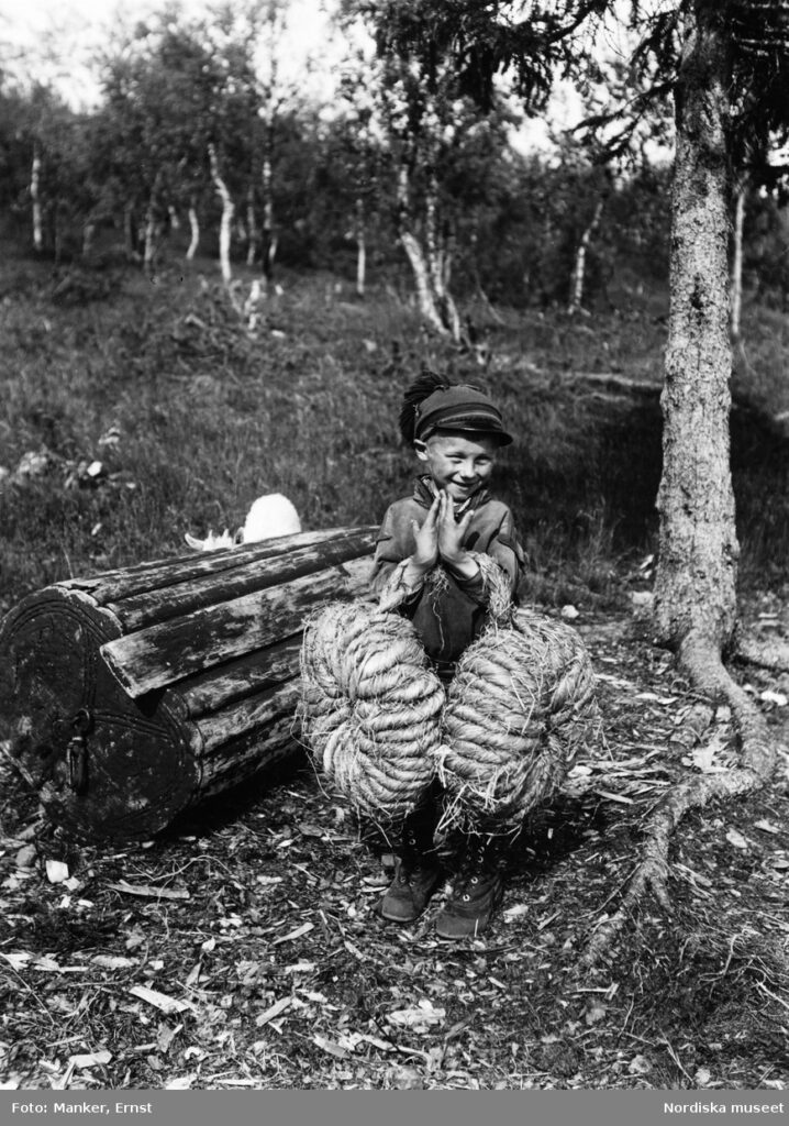 a black and white photo of a Sami boy holding two rolls of "shoe hay" standing next to an overturned wooden sledge.