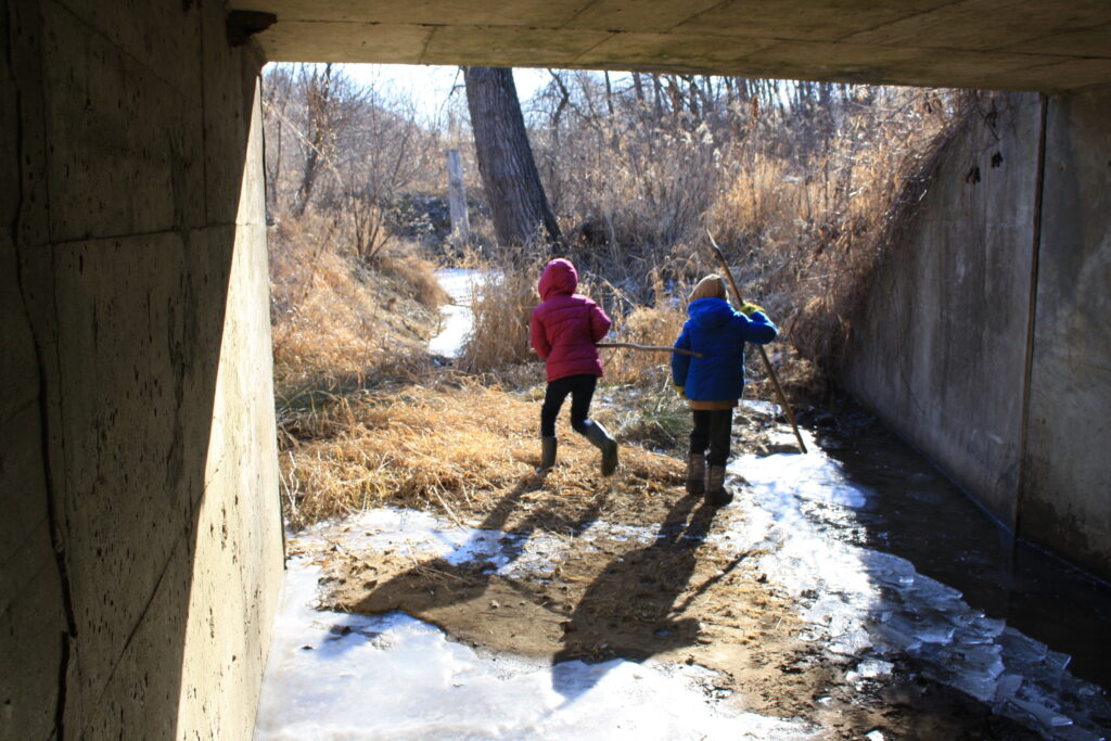 two outdoor children with walking sticks at the mouth of a tunnel with a creek and a woods.