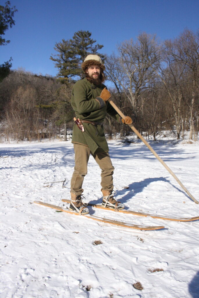 man in a green tunic with wooden medieval0-style skis and a long ski pole