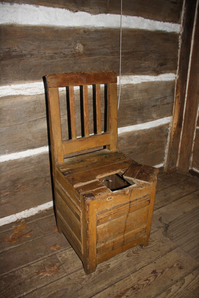 a wooden hay chair for storing hay to insulate footwear.  It has a hole on the seat, sitting in front of a log house wall.