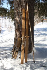 a pair of kid skis and a pair of adult wooden skis along with wooden ski poles leaning up against a tree trunk in the snow